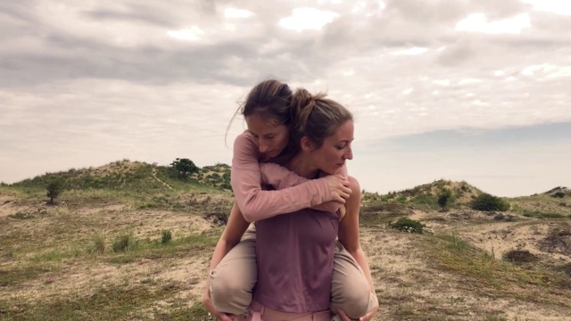 a woman carries another woman across grassy sand dunes