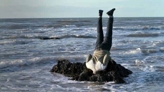 a man does a headstand on a small cluster of rocks near the ocean shore, head obscured