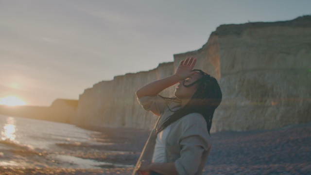 a light skinned woman with dark hair leans back gently, hand to forehead, with a sheer rock cliffside in the background