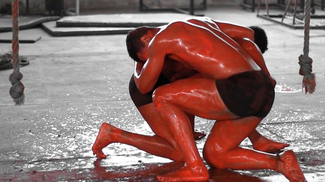 two men, bodies covered in a wet, red substance kneel in embrace