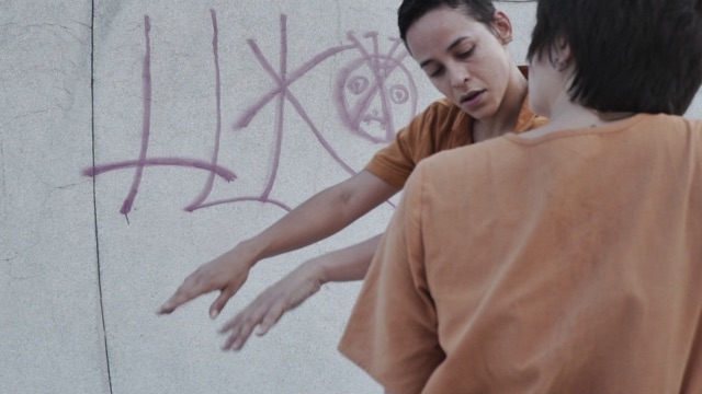 two people in pale orange shirts dance in front of a cement wall with simple graffiti