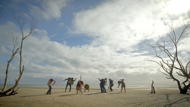 seven dancers gesticulate between two dead trees on a flat field of sand