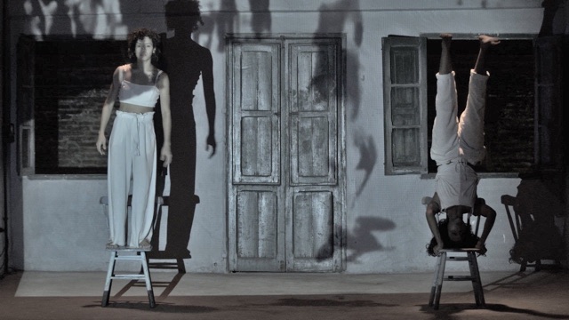 two performers in white on chairs - one standing on their feet, one standing on their head