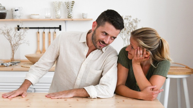 a man stands at a table, leaning heavily toward a woman who smiles in amusement