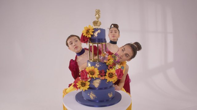 three young female dancers in fancy dresses examine an elaborate tiered cake