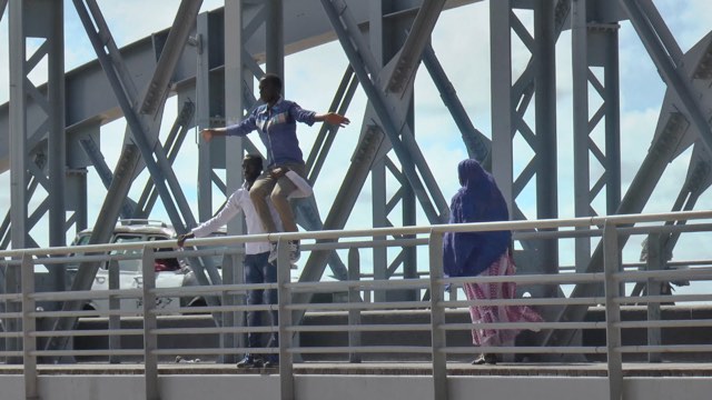 a dark skinned man sits on the shoulder of another man, who holds the railing of the bridge they stand on