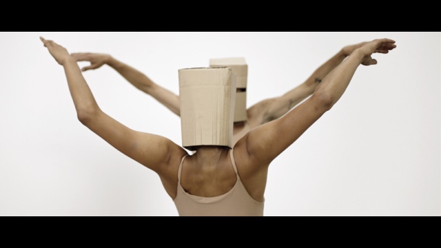 two dancers with square cardboard masks over their heads extend their arms out in a graceful, birdlike manner