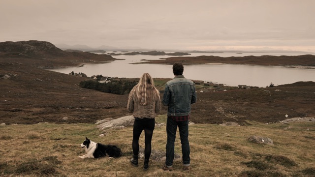 two people stand on a hilltop looking at a coastline with their dog beside them
