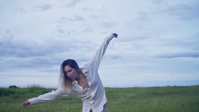 a woman wearing a loose white shirt extends her arms wide, leaning forward, in a grassy field with overcast skies above