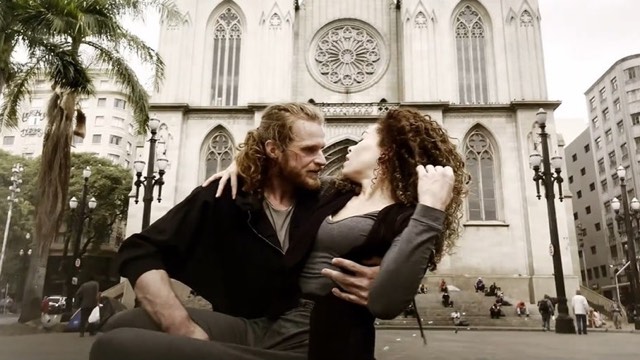 a long haired man lifts a curly haired woman in the courtyard of a large cathedral building