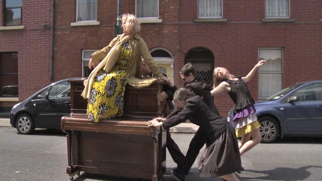 costumed dancers push an upright piano on wheels down a street while a woman in a yellow dress sits atop it