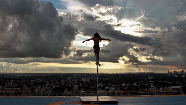 a woman balances atop a pole with the sun setting over a dense urban area in the background