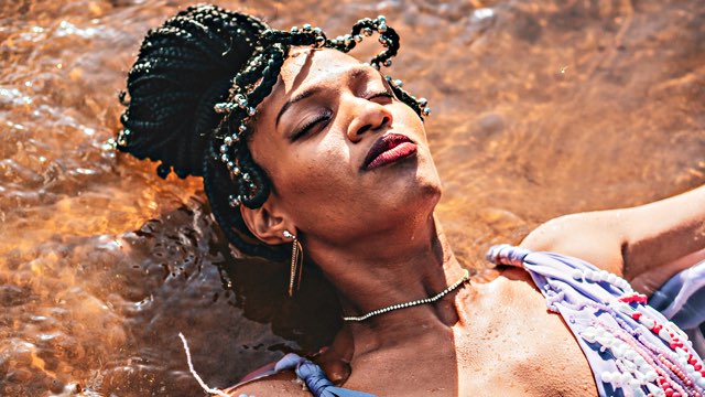 a person with braided hair and a dress lies on their back in shallow water