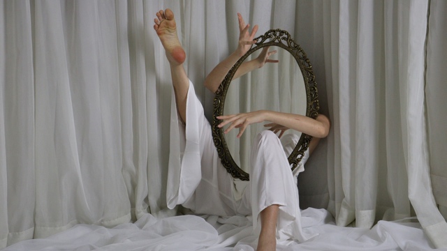 two arms and two legs protrude from behind a large white curtain - the arms holding a mirror