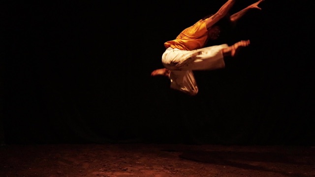 a dancer in an orange shirt and white pants leaps acrobatically across a dark room