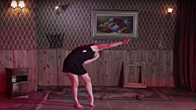 a light skinned woman in a black dress bends to the side in a sparsely decorated room