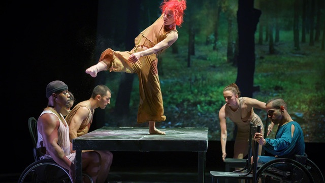 a woman with bright red hair dances atop a table attended by several dancers - some in wheelchairs