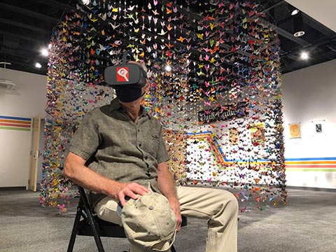 a man sits in a chair watching a dance film in a virtual reality headset; behind him is a wall of paper cranes