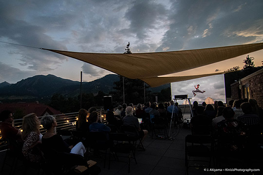 an audience watches a dance film on a large projection screen at dusk with mountains in the background