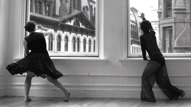 two dancers in black dresses dance facing away from the camera and looking out large windows out into a dense city