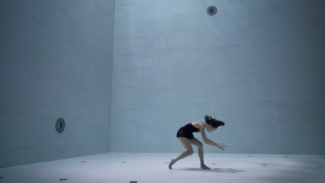 a female dancer stands underwater at the bottom of a deep swimming pool