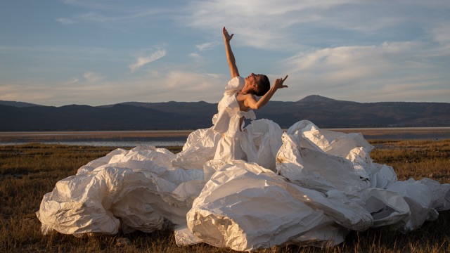 a dancer in a paper dress that unfolds around her reaches one hand toward the sky
