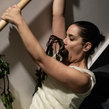 a light skinned woman climbs down a staircase bannister with a root vegetable in her mouth