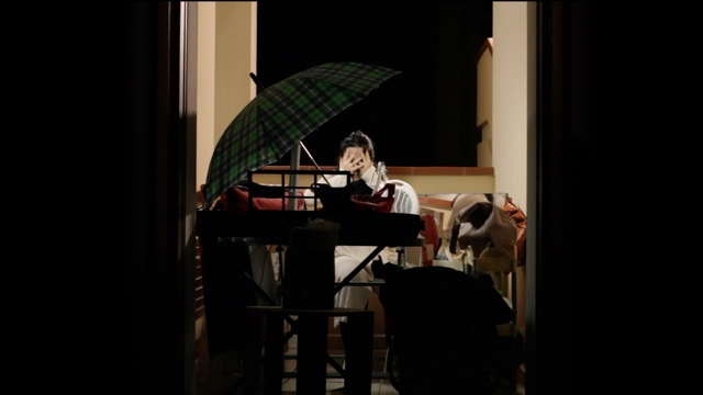 a dancer sits at a crowded desk with her hands over her eyes, with an umbrella in the foreground