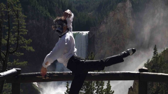 a lean dancer in black dress pants, a formal white shirt, and an elaborate mask strikes a pose in front of a huge waterfall