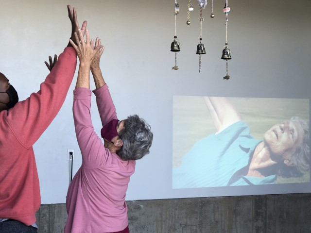 a dark skinned man and grey haired woman reach upwards in unison with hands touching