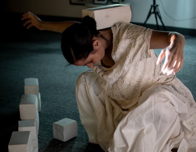 a light skinned woman in a white dress balances a block on the back of her neck with other blocks positioned in front of her