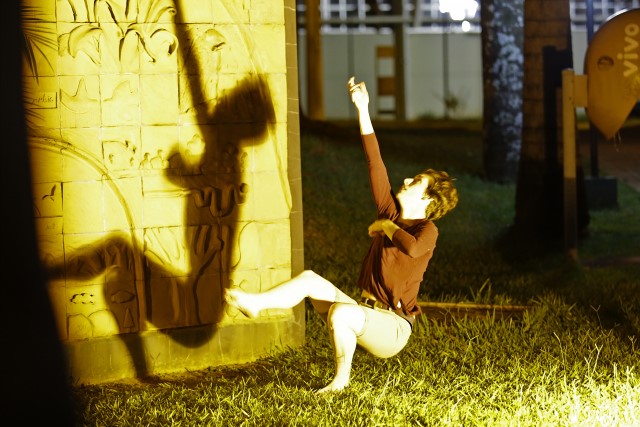 a short haired woman squats and balances on one foot as her shadow is projected onto a stone carving behind her