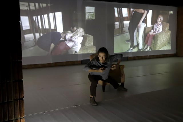 a brown haired woman leans forward in front of a stuffed chair while films are projected onto the wall behind her
