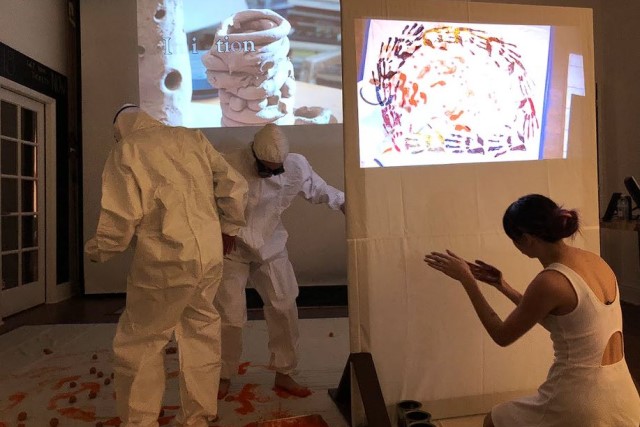 two people in white painting suits step in paint while a dancer in a white dress looks at her paint-covered hands