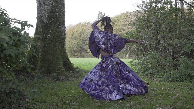 a woman with dreadlocks wearing a purple dress covered in circular shapes extends her right arm while facing a clearing in a forest