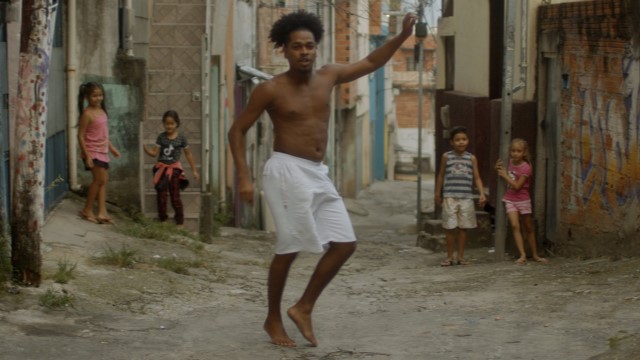 a dark skinned man dances shirtless in an alley while four children smile and watch