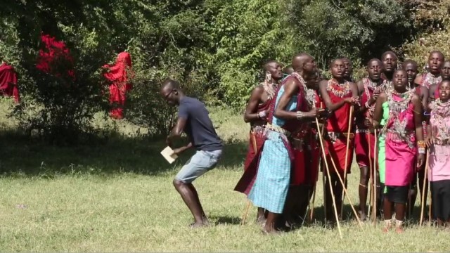 a group of Maasai people holding long sticks sing in unison while a dark skinned man crouches to their right