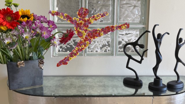 a small glass table in the entryway of a home displays three dancing figurines, a vase of flowers, and a miniature leaping dancer composed of flowers