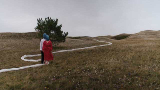 two people with towels wrapped around their heads and faces stand in a meadow next to a long and winding line of white fabric
