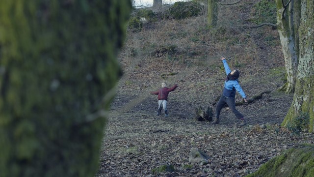 a tree partially obscures the foreground; behind it we see an adult and a child striking a pose in the woods