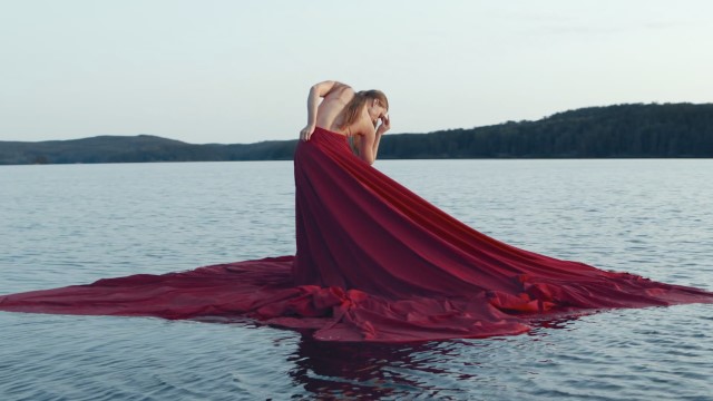 a light skinned woman in an extremely long red dress seemingly stands on the surface of a lake