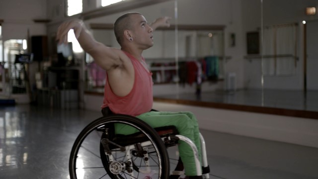 a man in a wheelchair gazes upward in a dance studio, arms outstretched