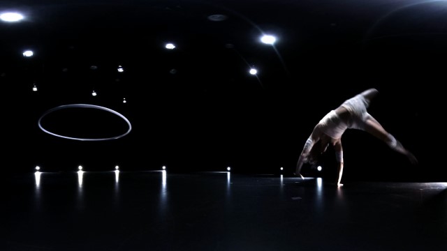 a large dark stage is illuminated by points and rings of light while a dancer cartwheels toward a suspended hoop