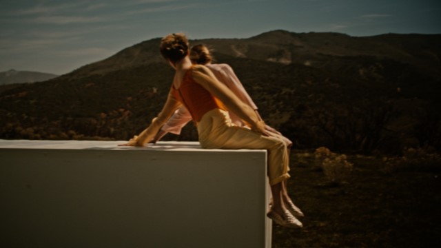 two women lean back on a large rectangular object with foothills in the background