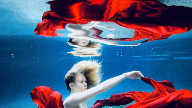 a light skinned woman in a long red dress floats just below the surface in clear water, eyes open