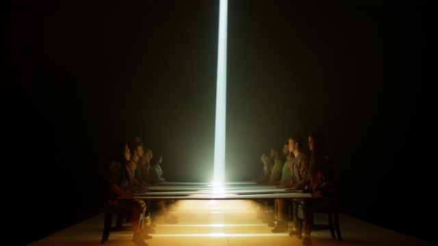 five people sit on each side of a long table in a dark room illuminated by a bright column of light