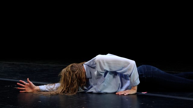 a person with long brown hair lays face down on a black stage floor, right arm outstretched