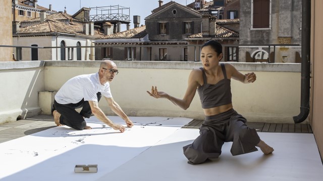 an Asian woman and white man dance while kneeling on white panels atop an urban roof