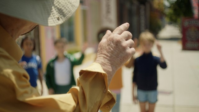 an old man waves to four children outdoors on a sidewalk