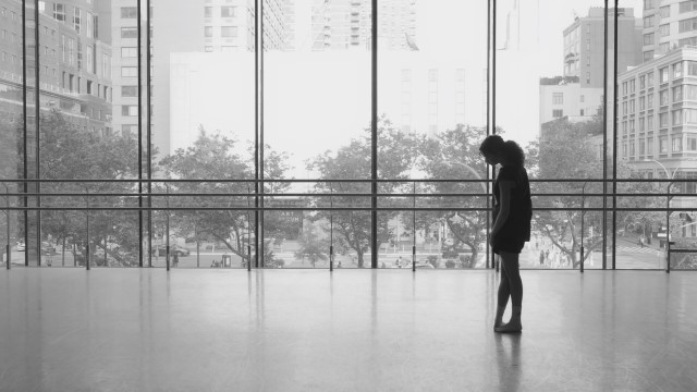 a black and white photo of a long haired dancer looking out huge glass windows to a busy city street